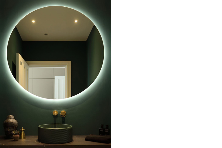 Backlit round bathroom mirror on dark green walls. Sit on sink with brass fittings.