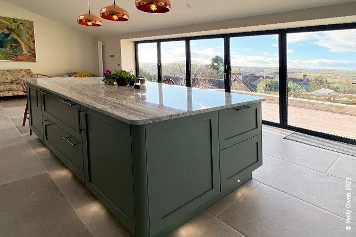 Large bifold windows look over the stunning Cotswolds views. A large kitchen island is under three copper lights.